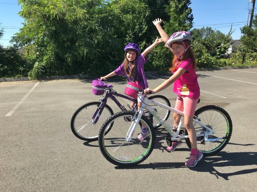 Two children wearing helmets rode bicycles in a sunny parking lot, one of them raising an arm in the air.