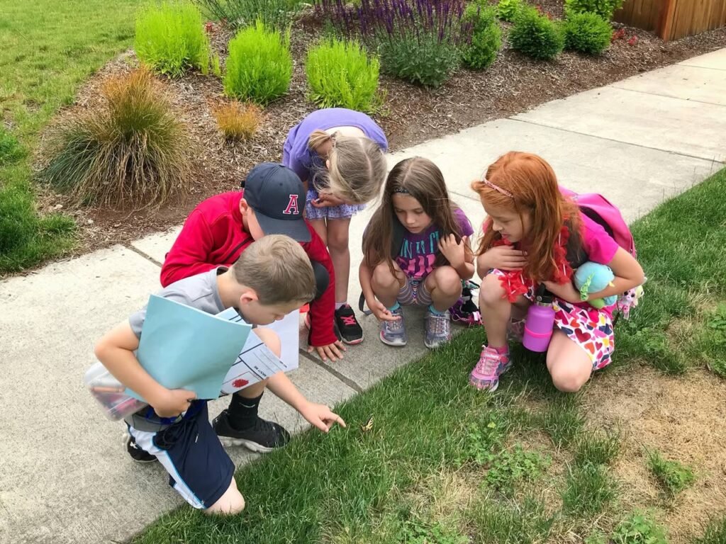 A group of children crouching near a garden path, examining plants. One child carries a potted plant in a blue container, set against a backdrop of ornamental grasses.