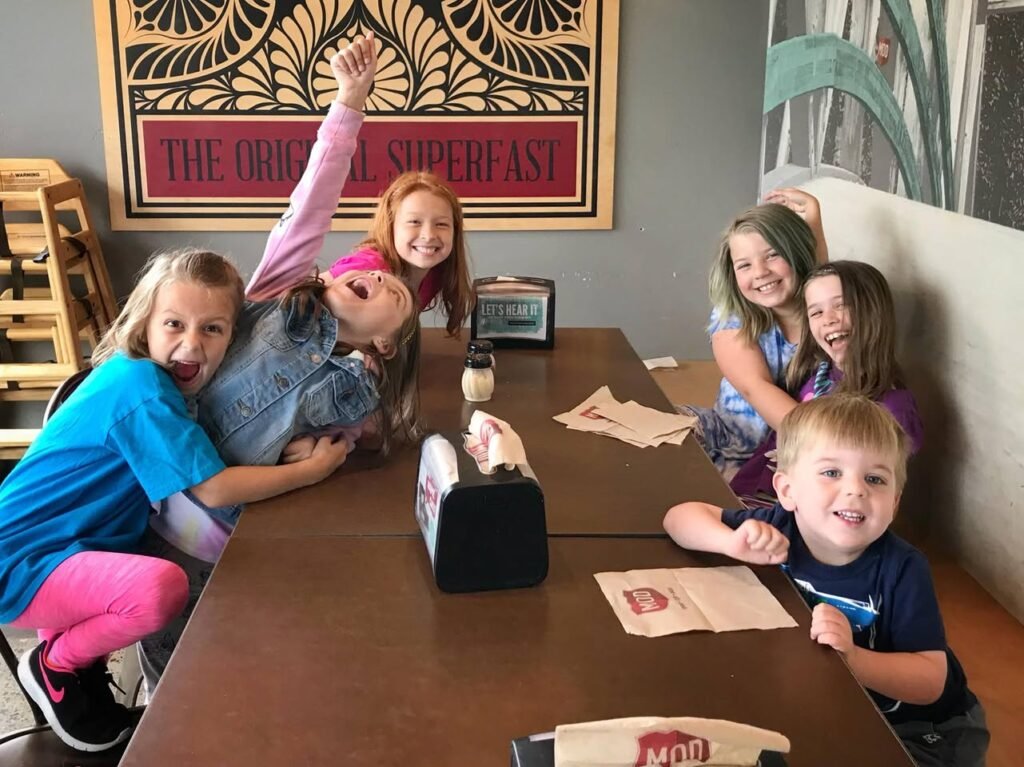 A group of children are gathered around a table in a booth, posing playfully for a photo. One child is dressed in a colorful outfit, while others seem to be enjoying a meal.