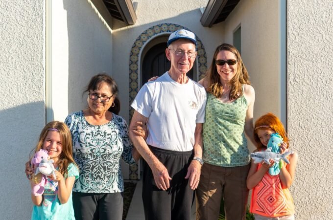 A family of five posing for a photo in front of an adorable building. The two young girls are each holding a stuffed animal.