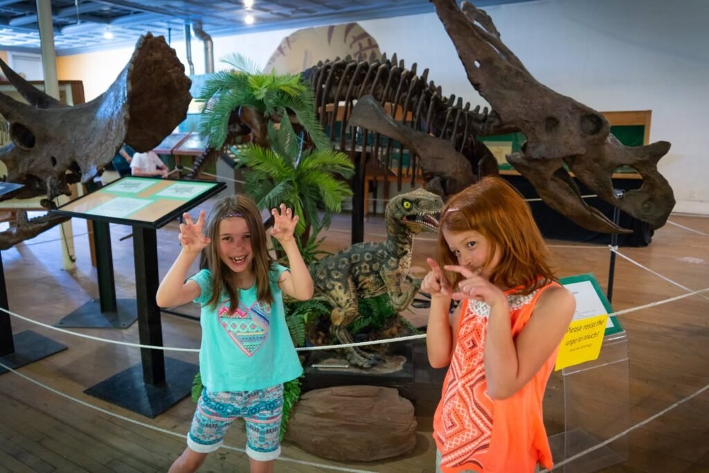 Two kids posing playfully in front of dinosaur skeletons and fossils at a natural history museum, surrounded by educational displays.