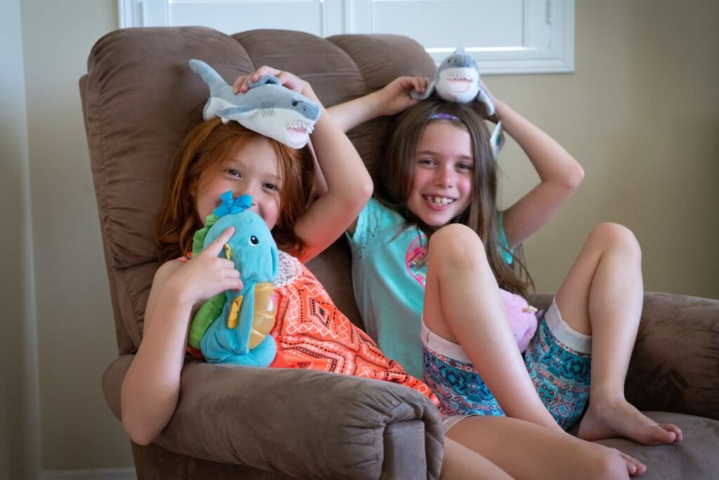 Two children sit in a brown armchair wearing plush shark hats while holding stuffed toys.
