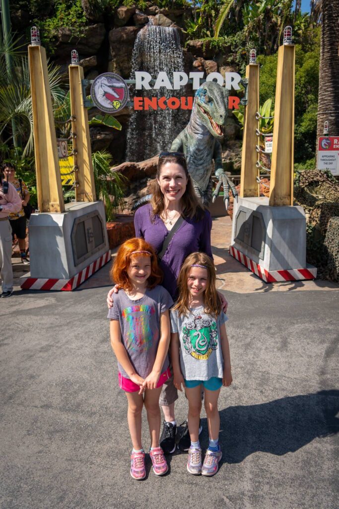 A woman and two children standing in front of the sign for Universal's Raptor Encounter attraction. The scene features a water feature and tropical landscaping.