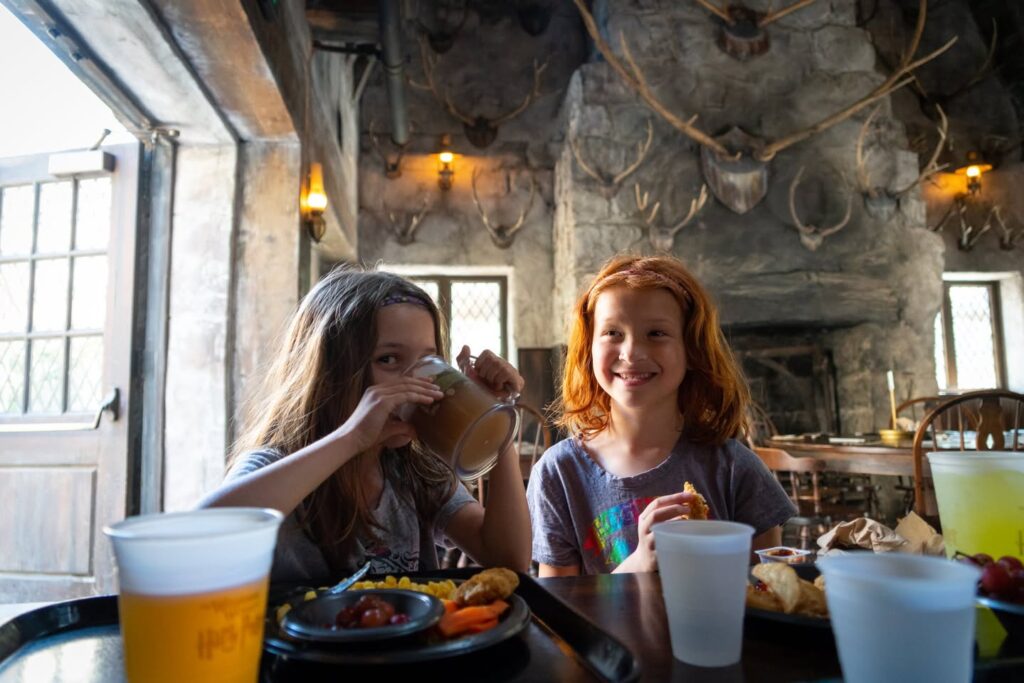 Two visitors, one with red hair, sit at a table in Hagrid's Hut at the Wizarding World of Harry Potter, holding glasses of Butterbeer near a table laden with snacks.