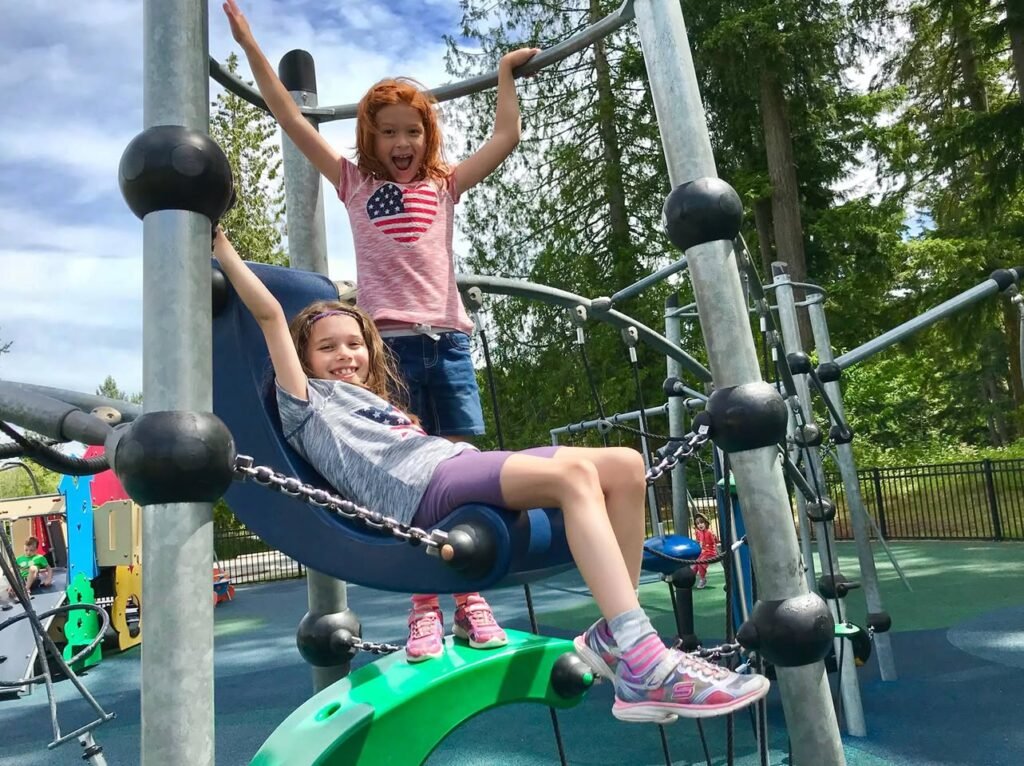 Two people playing on a playground structure with one seated on a swing and the other climbing. The background features trees and a blue sky.