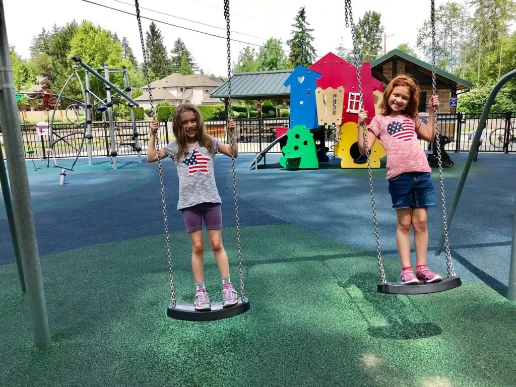 A photo collage showing children playing on swings at a colorful playground. They are wearing casual summer clothes and standing on green safety surfacing.
