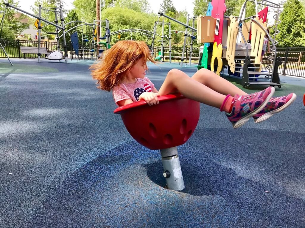 A young child with red hair enjoys a red spinning saucer at an outdoor playground. Colorful play structures and green trees create a vibrant summer backdrop.