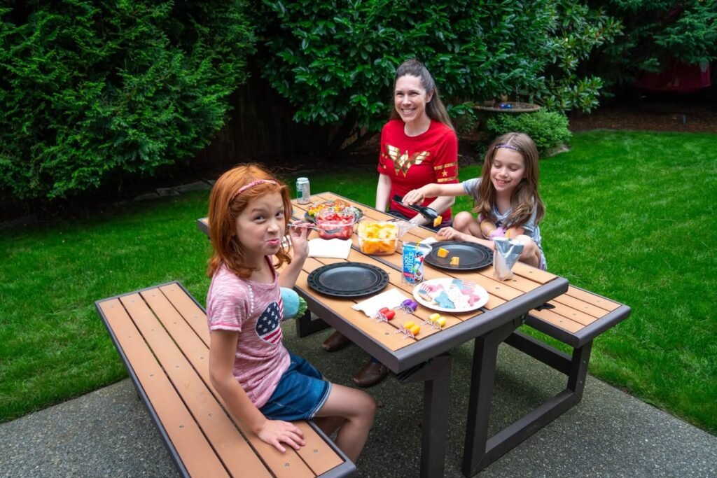 A family gathers around a wooden picnic table for a casual outdoor meal. Green foliage and a well-kept lawn surround the patio setting.