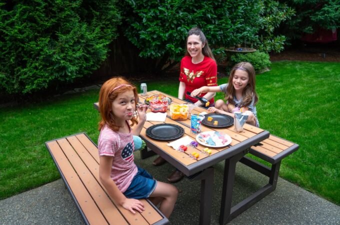 A family gathers around a wooden picnic table for a casual outdoor meal. Green foliage and a well-kept lawn surround the patio setting.