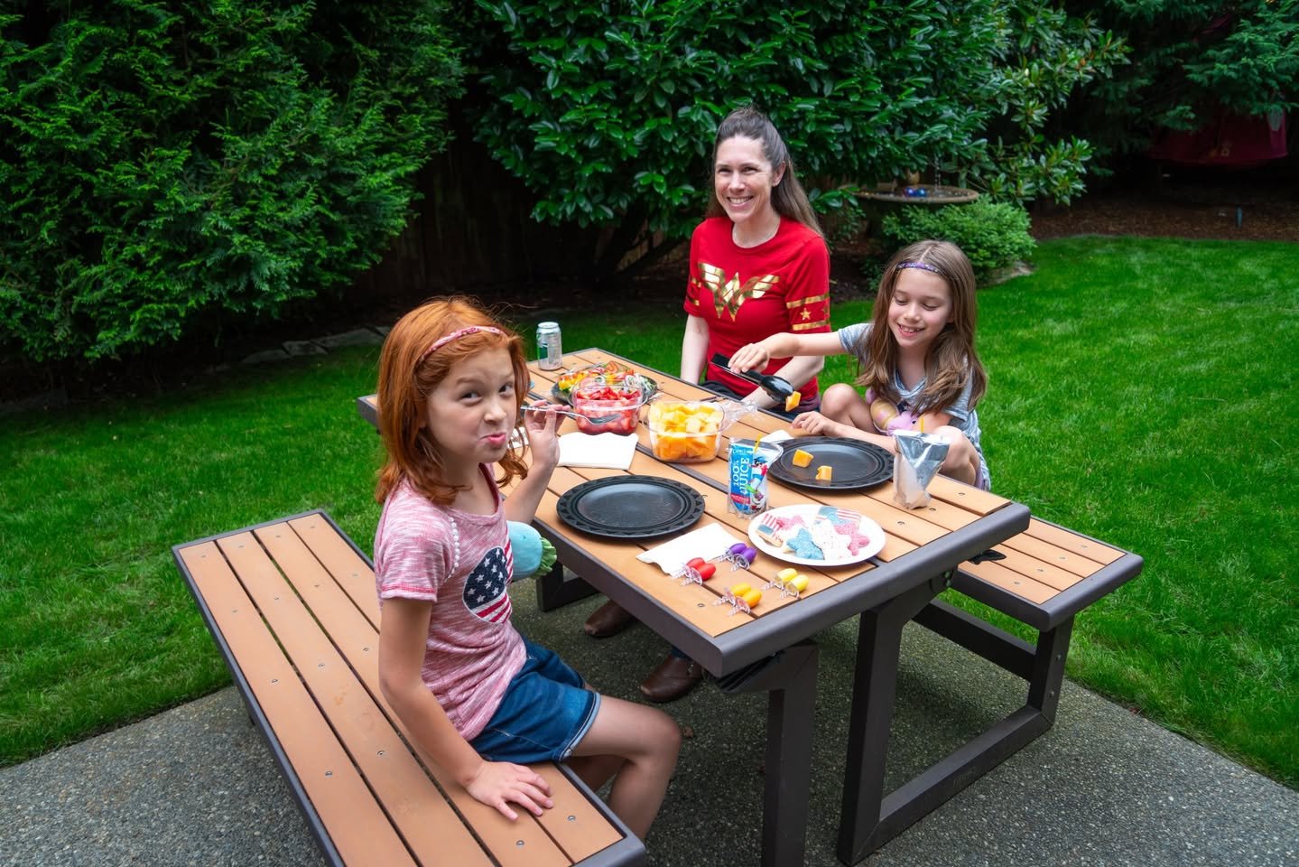 A family gathers around a wooden picnic table for a casual outdoor meal. Green foliage and a well-kept lawn surround the patio setting.