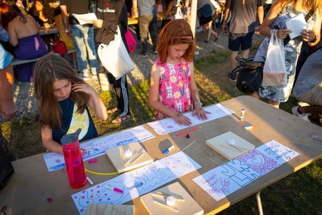 Children participate in fabric dyeing activities on a table with paper designs during a sunny daytime outdoor festival.