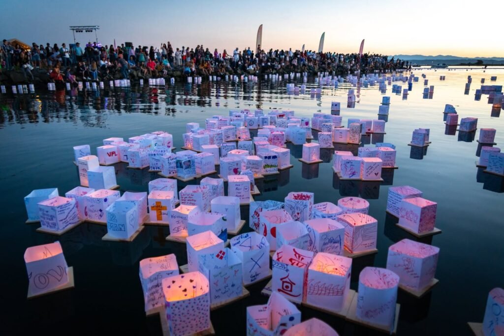Hundreds of illuminated paper lanterns float on the water at dusk while a crowd watches from the shore during the Screaming Frog Festival.