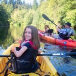 A person paddles a yellow kayak down a serene river, followed by others in an orange kayak through a rich, green forest.