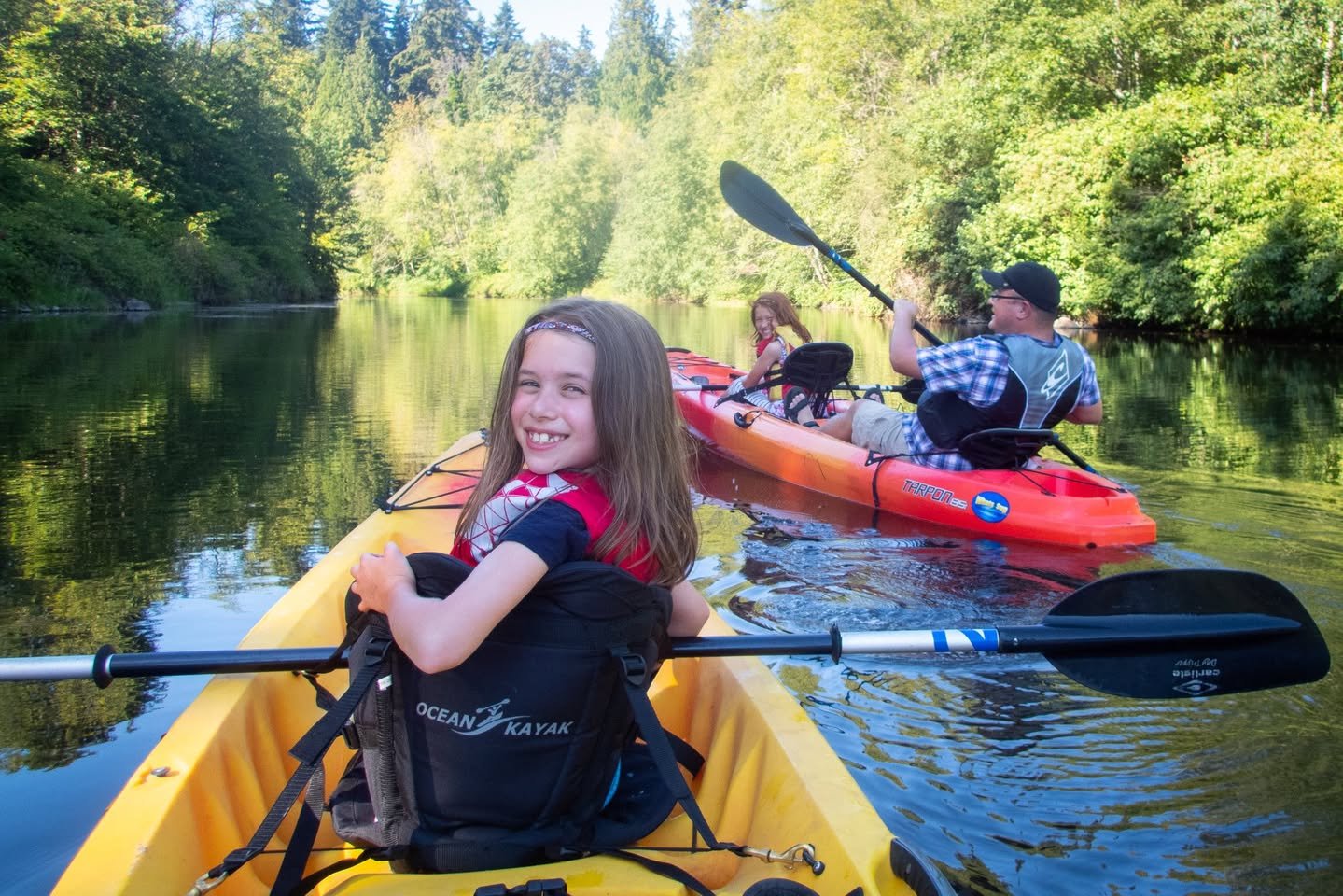A person paddles a yellow kayak down a serene river, followed by others in an orange kayak through a rich, green forest.