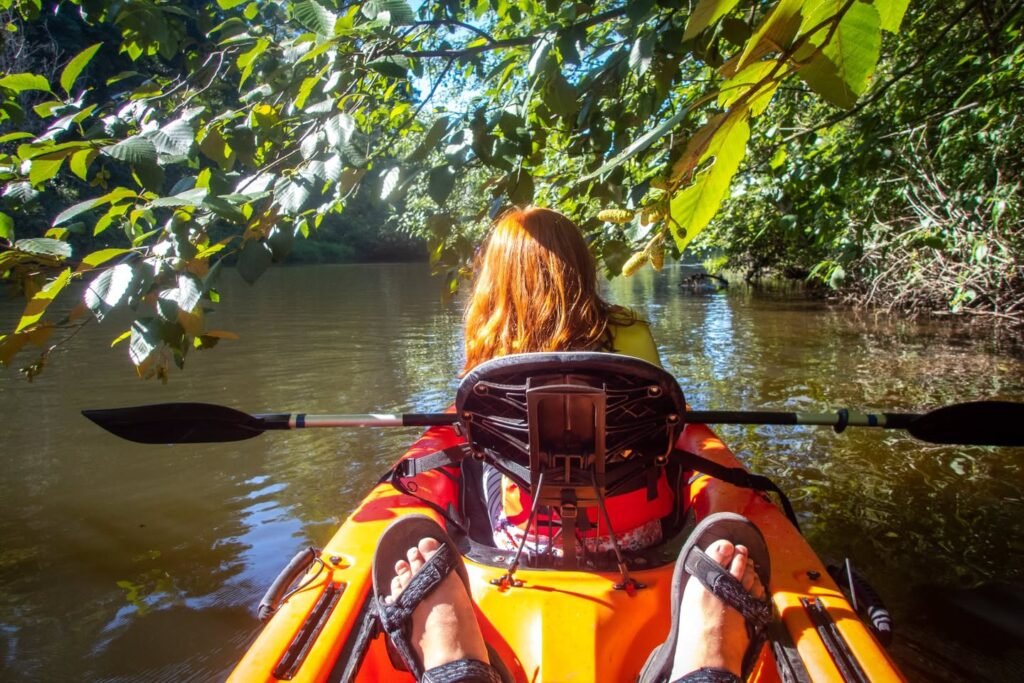 View from a yellow kayak paddling along a calm river surrounded by dense greenery. Sunlight filters through leaves as the paddle dips into the water.