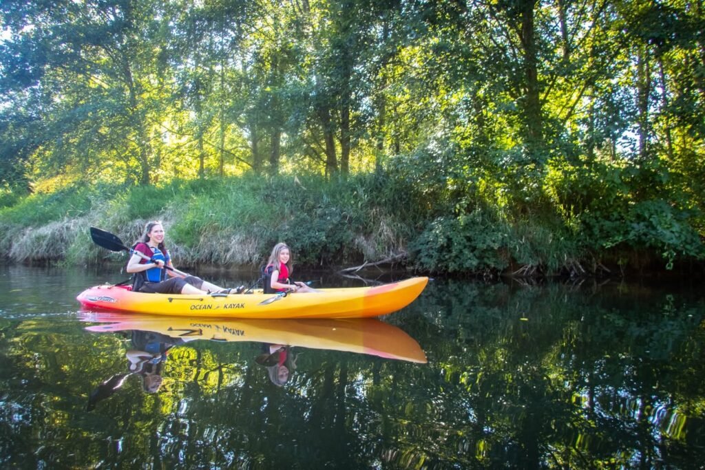 A bright yellow kayak glides along a calm river reflecting the lush green trees.