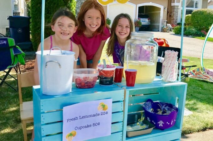 A colorful homemade lemonade stand made from painted wooden crates. A yellow lemonade banner hangs above a family enjoying the sunny day.