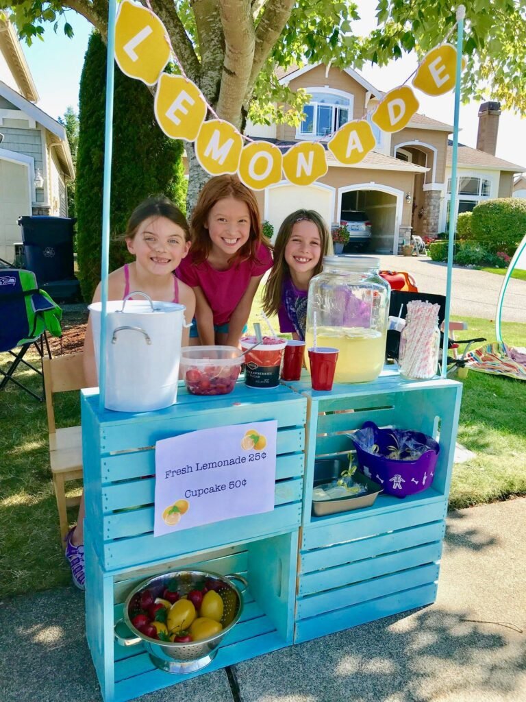 A colorful homemade lemonade stand made from painted wooden crates. A yellow lemonade banner hangs above a family enjoying the sunny day.