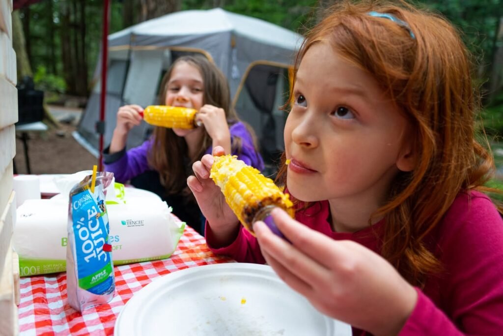 Two children eating fresh corn on the cob while camping outdoors.