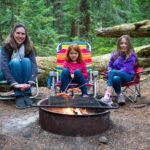 Three people sitting in colorful camping chairs around a campfire in a lush green forest.