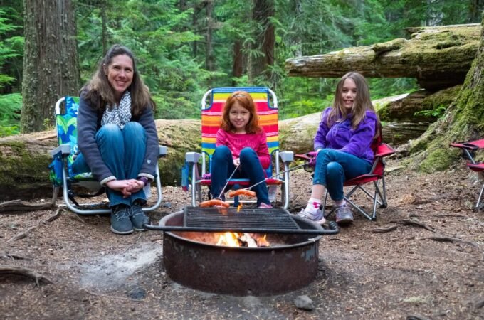 Three people sitting in colorful camping chairs around a campfire in a lush green forest.