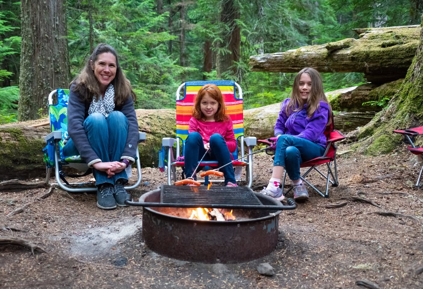 Three people sitting in colorful camping chairs around a campfire in a lush green forest.