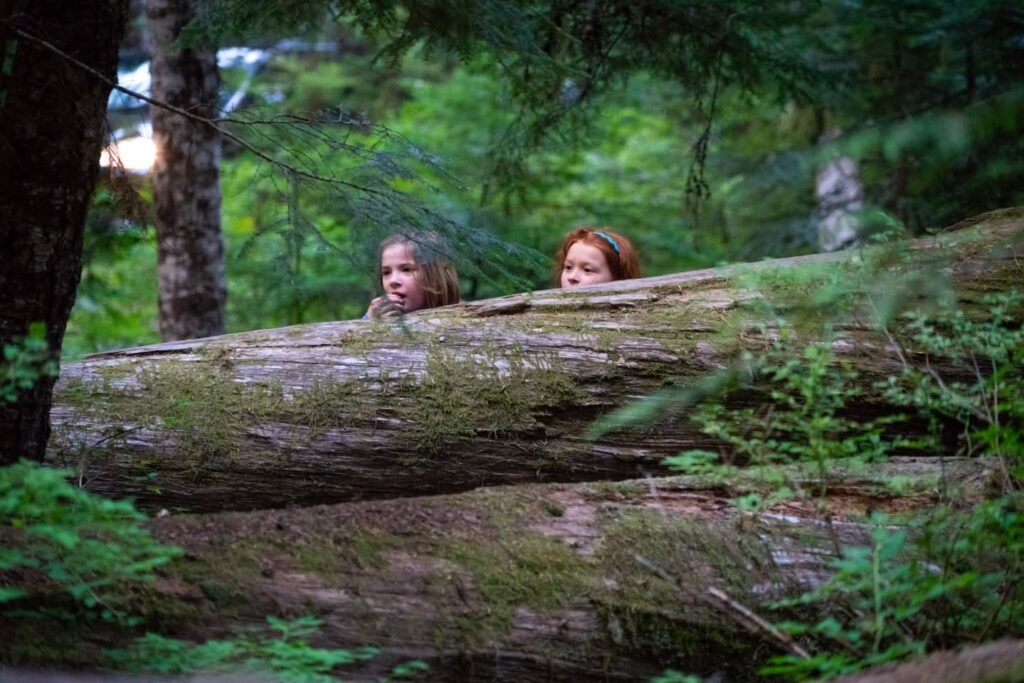 Children peeking from behind a mossy log in a lush green forest.