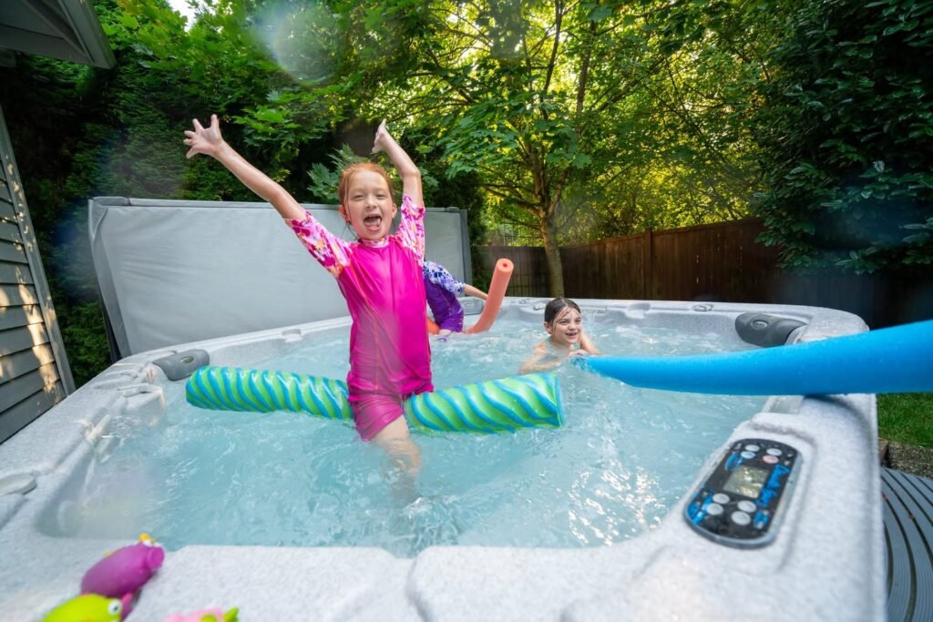 A child celebrates with arms raised in a backyard hot tub surrounded by greenery.