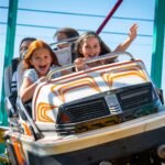 Smiling riders enjoying a car shaped theme park ride under a bright blue sky.