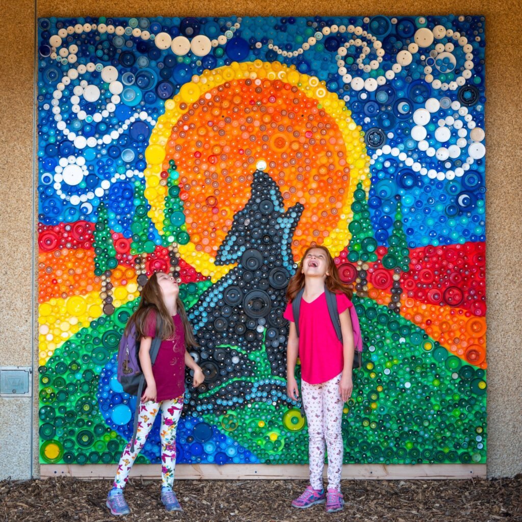 Two young people standing in front of a vibrant mosaic mural featuring a bear and a large orange sun.