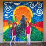 Two children posing in front of a colorful handcrafted mosaic art piece featuring a sun and celestial swirls.