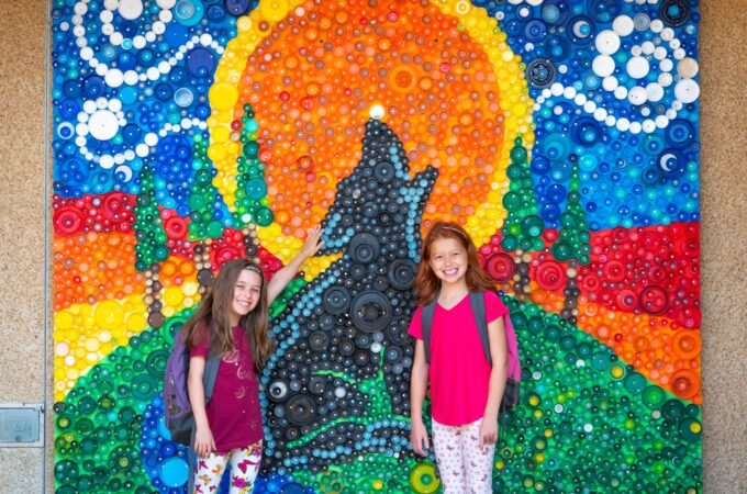 Two children posing in front of a colorful handcrafted mosaic art piece featuring a sun and celestial swirls.
