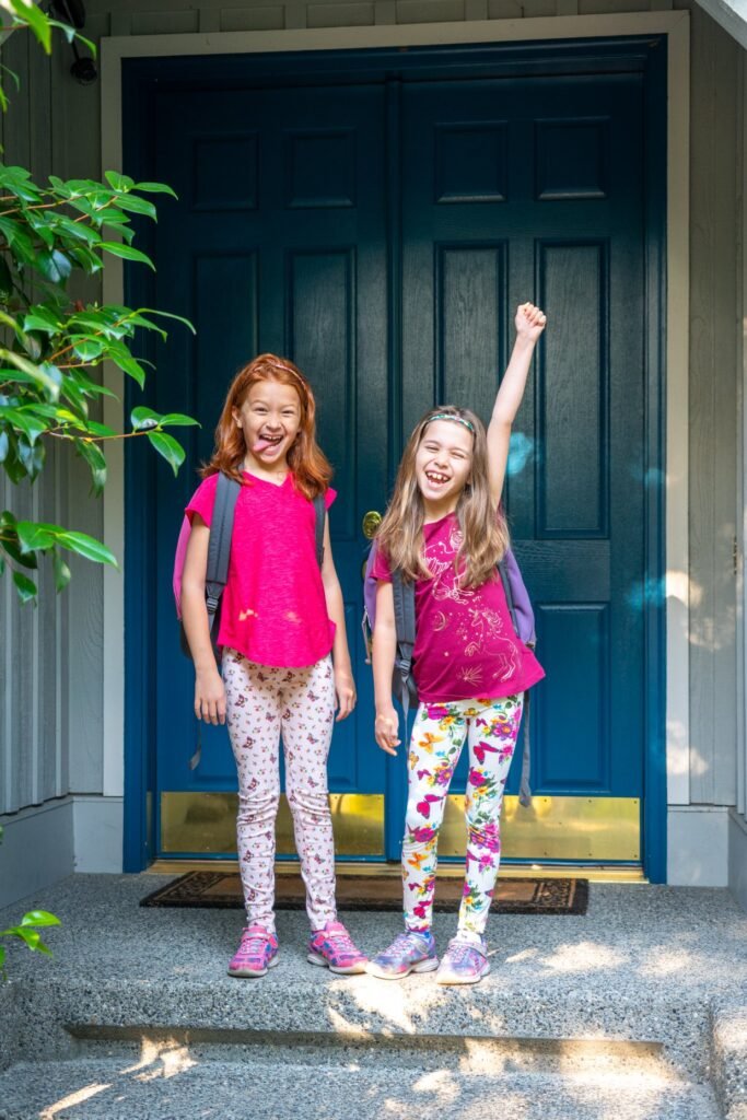 Two children stand cheerfully on steps outside a blue framed doorway, wearing colorful outfits and backpacks.