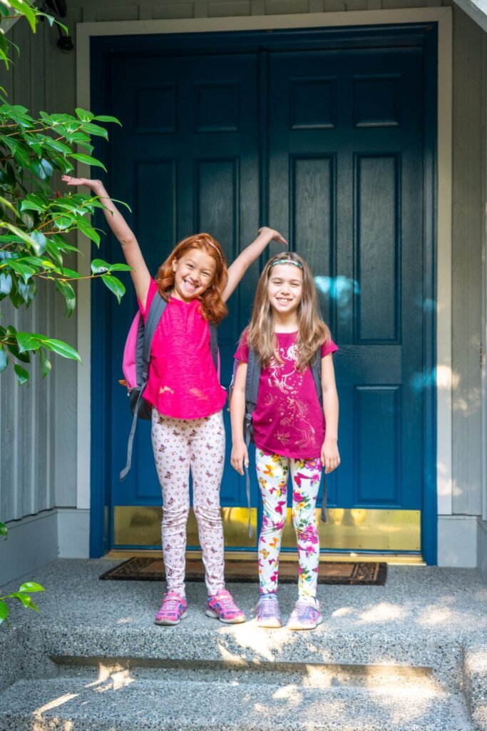 Two girls stand together in front of a teal entryway, one with arms raised. They are wearing vibrant floral and patterned outfits.