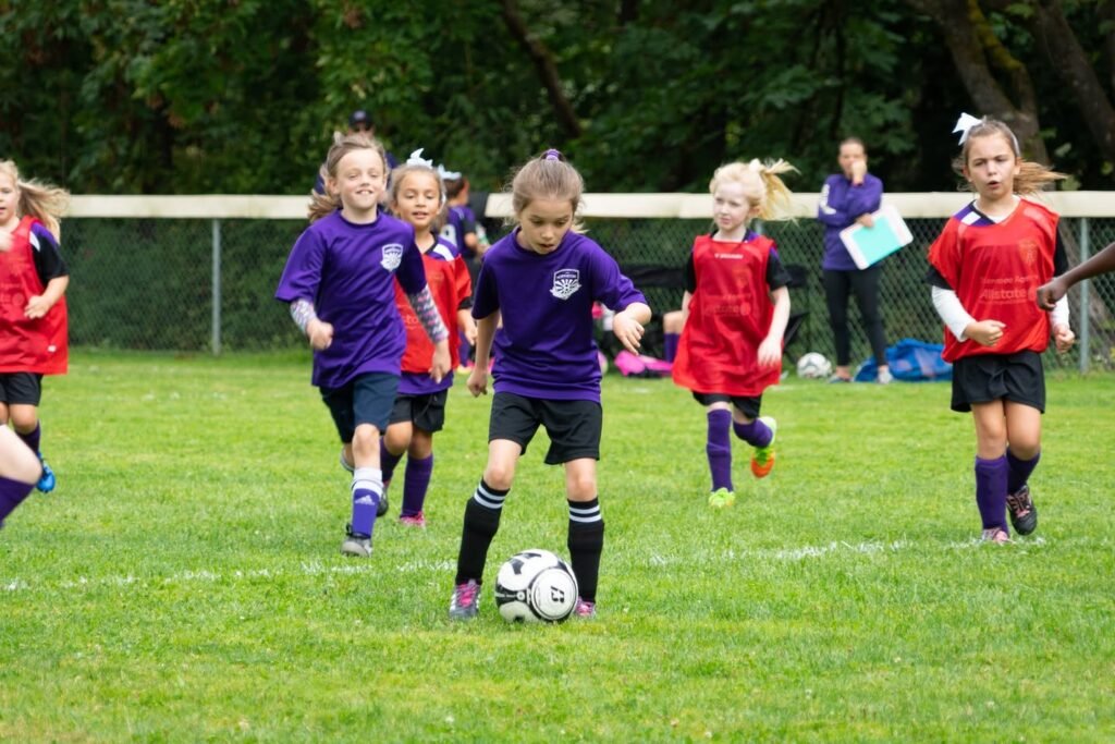 Children in purple and red uniforms play soccer on a grassy field. A player in purple dribbles the ball while teammates and opponents run nearby.