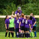 A youth soccer team in purple jerseys huddles together with a coach on a grass field with lush green trees in the background.