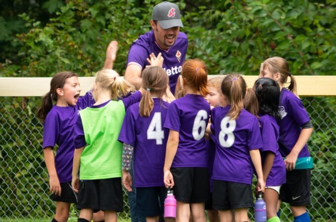 A youth soccer team in purple jerseys huddles together with a coach on a grass field with lush green trees in the background.