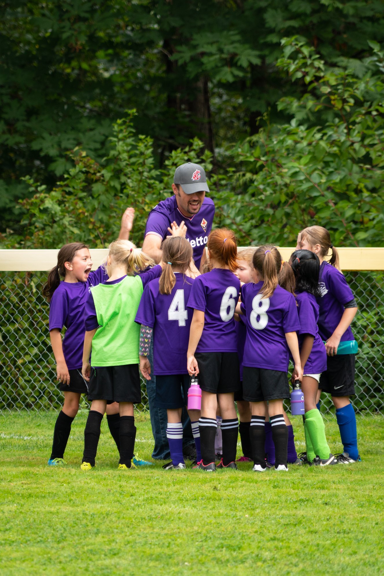 A youth soccer team in purple jerseys huddles together with a coach on a grass field with lush green trees in the background.