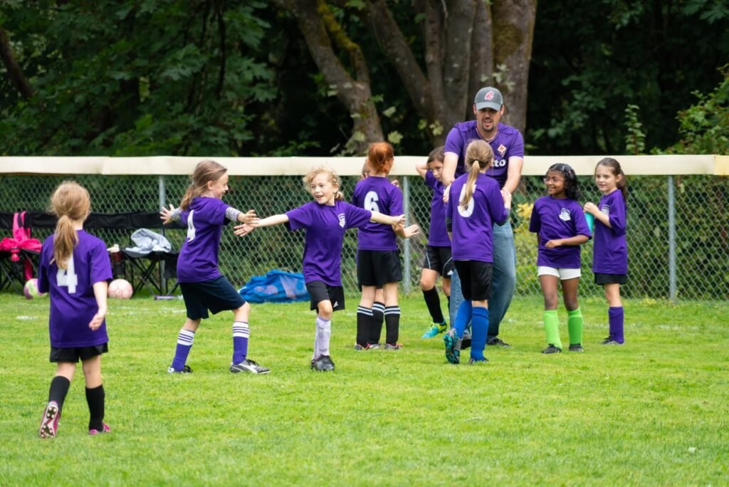 Group of children in purple jerseys playing soccer on a grassy field. An adult in purple supervises nearby.