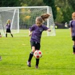 A young player in a purple uniform chasing a soccer ball on a sunny day.