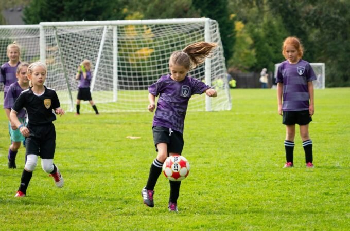 A young player in a purple uniform chasing a soccer ball on a sunny day.