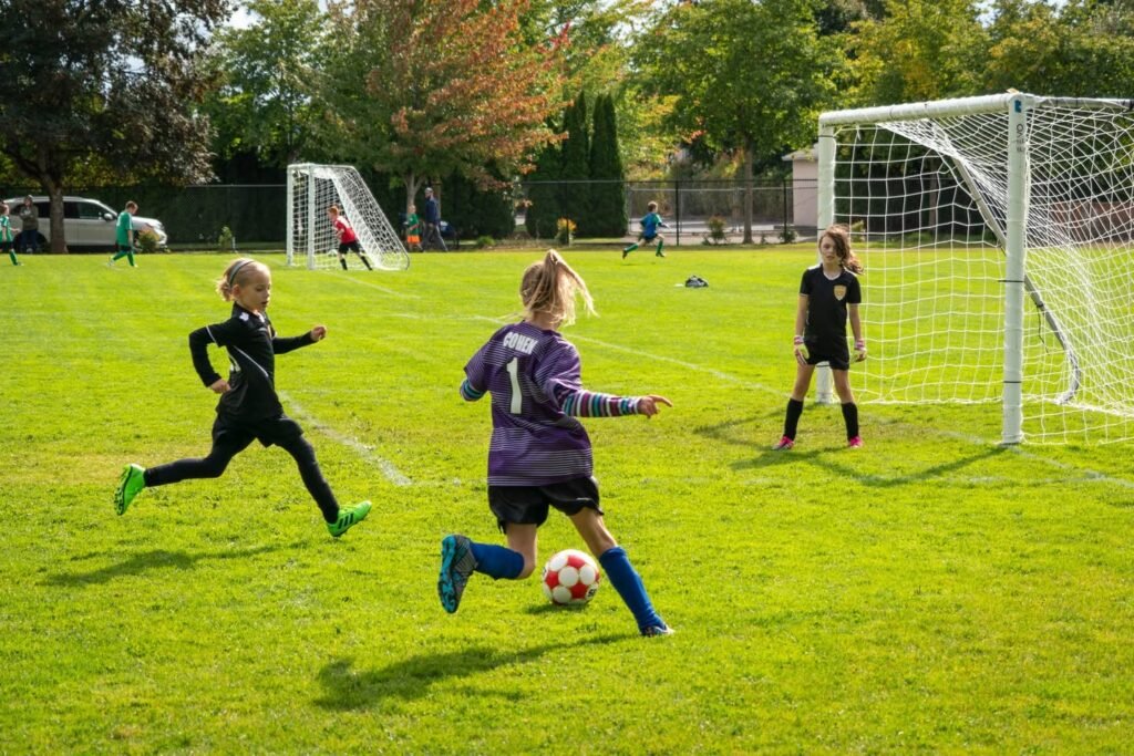 Four photos showing young children playing soccer on a green grass field. A player in purple kicks a ball while others watch near the goal.