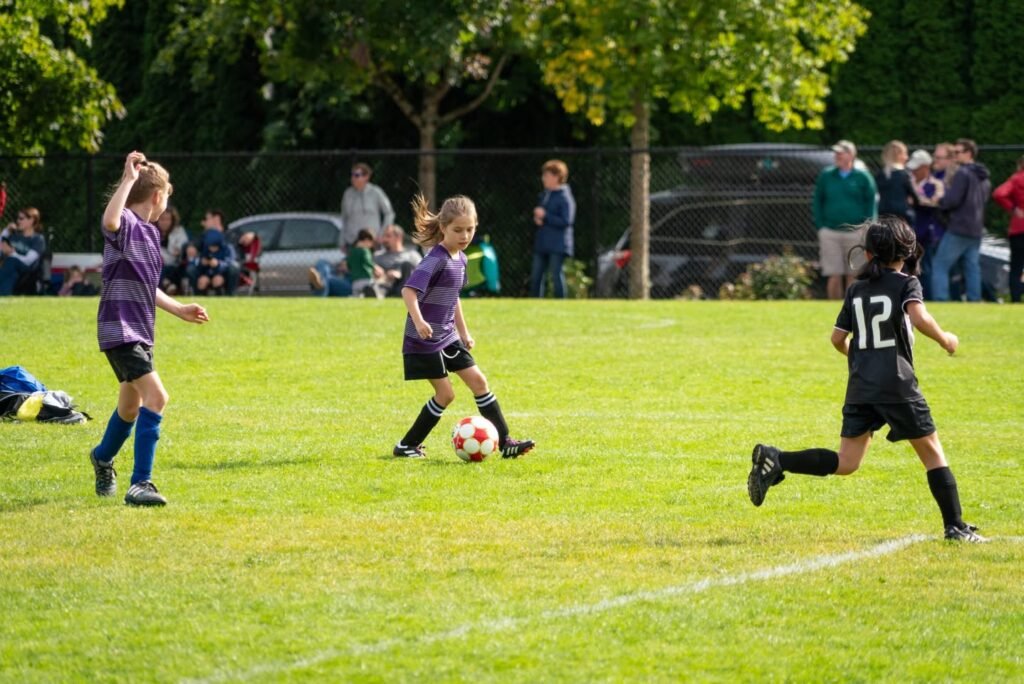 A young soccer player in a purple jersey dribbles a soccer ball on a grass field while chasing a defender in black.