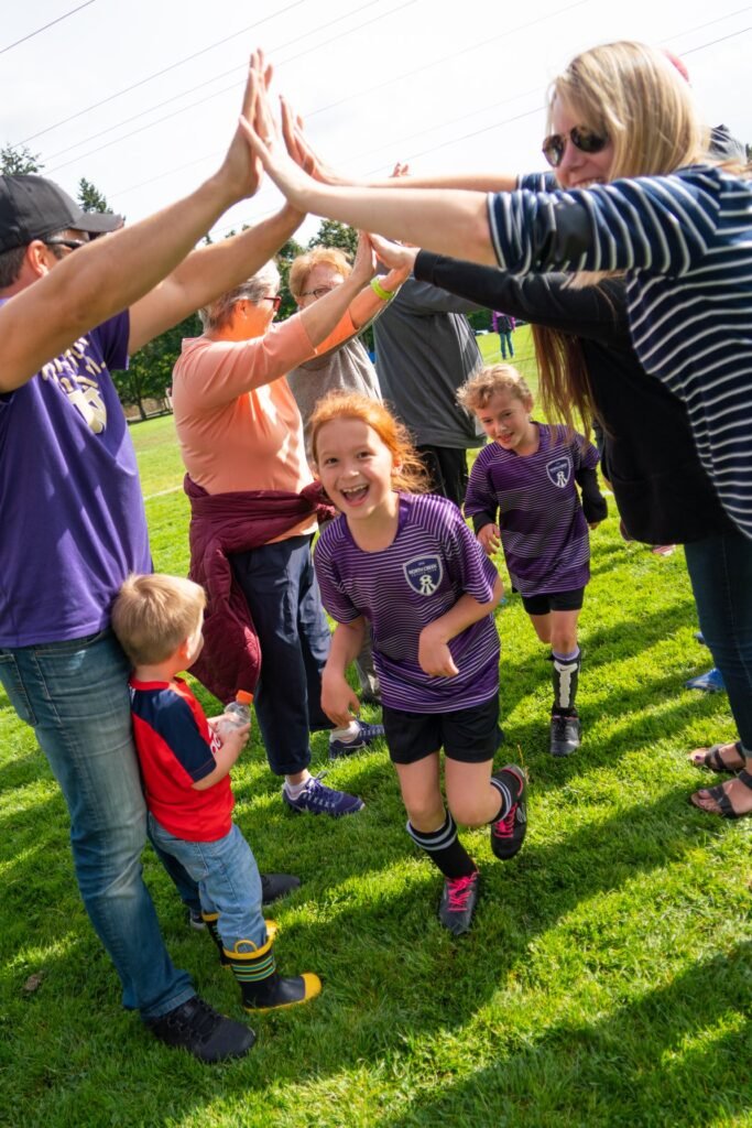A group of children in purple uniforms high-five adults at a sunny outdoor sports event.