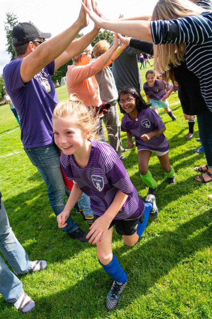 Children in purple soccer uniforms high-fiving adults on a grassy field, creating a joyful atmosphere.