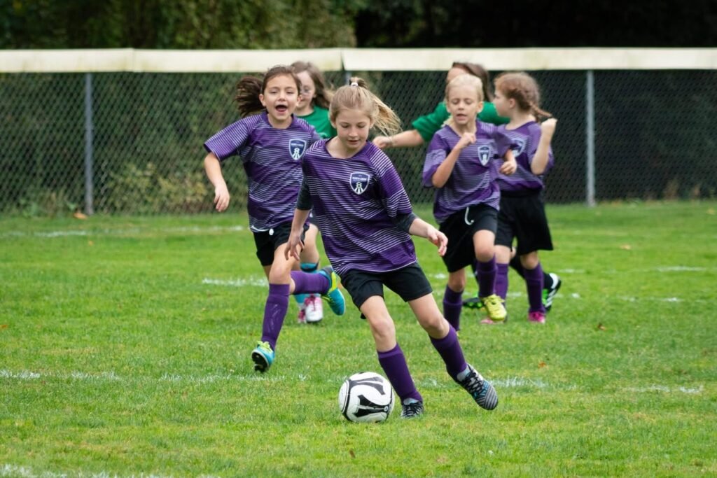 Junior soccer players in purple uniforms compete for the ball during an outdoor match on a green field.