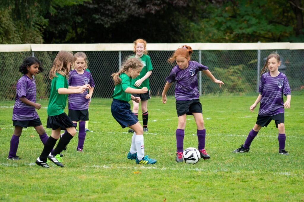 Young players in purple and green uniforms compete during an outdoor youth soccer game on a grass field.