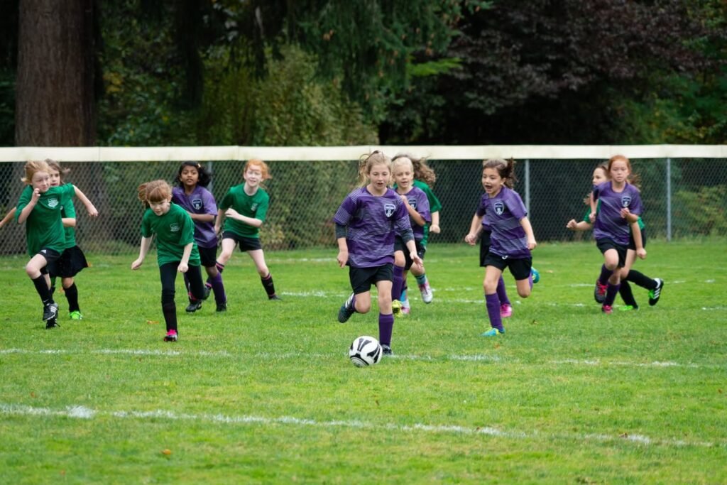 Young players in green and purple jerseys compete on a grassy soccer field. Trees and a fence line the background, capturing a lively outdoor sports event.