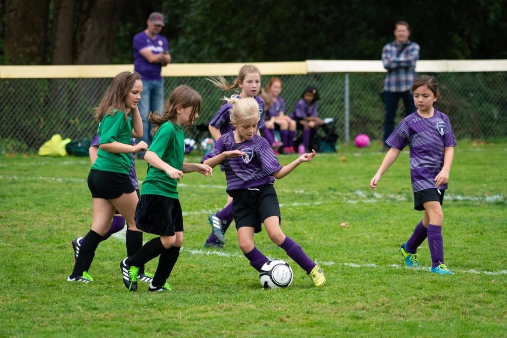 A youth soccer player in purple controls the ball during a game. Teammates and opponents in green and purple jerseys are visible on the grass field.