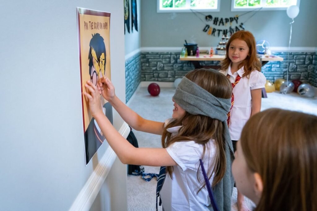 A child in a blindfold reaches out to touch a card on the wall at a birthday party. A banner in the background says Happy Birthday.